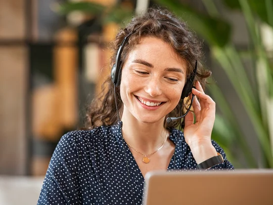Junge Frau sitzt lächelnd am Schreibtisch, arbeitet am Laptop und telefoniert gleichzeitig mit einem Kunden – typische Szene einer Service-Desk-Beraterin im Callcenter.
