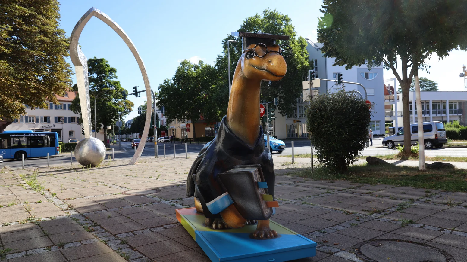 Maskottchen der Hochschule Worms, ein Dinosaurier mit Doktorhut, Brille und Büchern unter dem Arm, als Skulptur auf einem Gehweg in Worms, im Hintergrund eine Bushaltestelle und eine metallene Bogenskulptur.