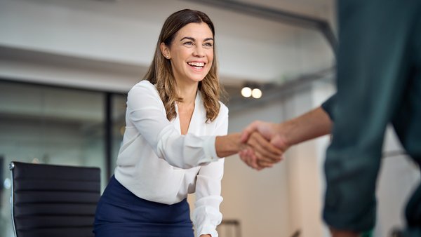 Lächelnde Geschäftsfrau, die mit einem Partner am Besprechungstisch im Stehen ein erfolgreiches Geschäft abschließt und ihm die Hand schüttelt.