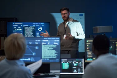 IT-Security-Spezialist erklärt einem Team Cybersecurity-Daten vor mehreren Monitoren.