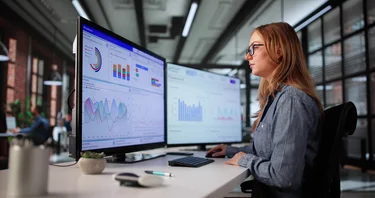 Frau analysiert Daten und Diagramme auf zwei Monitoren.