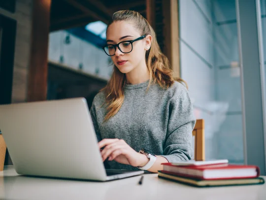 Interessierte Studentin informiert sich online über Studiengänge und Angebote der Hochschule. Über WLAN mit dem Internet verbunden, recherchiert die aufgeschlossene junge Frau mit Laptop und Brille gezielt nach passenden Studienmöglichkeiten.