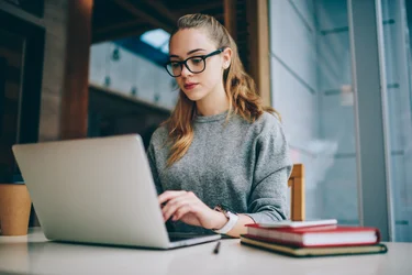 Interessierte Studentin informiert sich online über Studiengänge und Angebote der Hochschule. Über WLAN mit dem Internet verbunden, recherchiert die aufgeschlossene junge Frau mit Laptop und Brille gezielt nach passenden Studienmöglichkeiten.