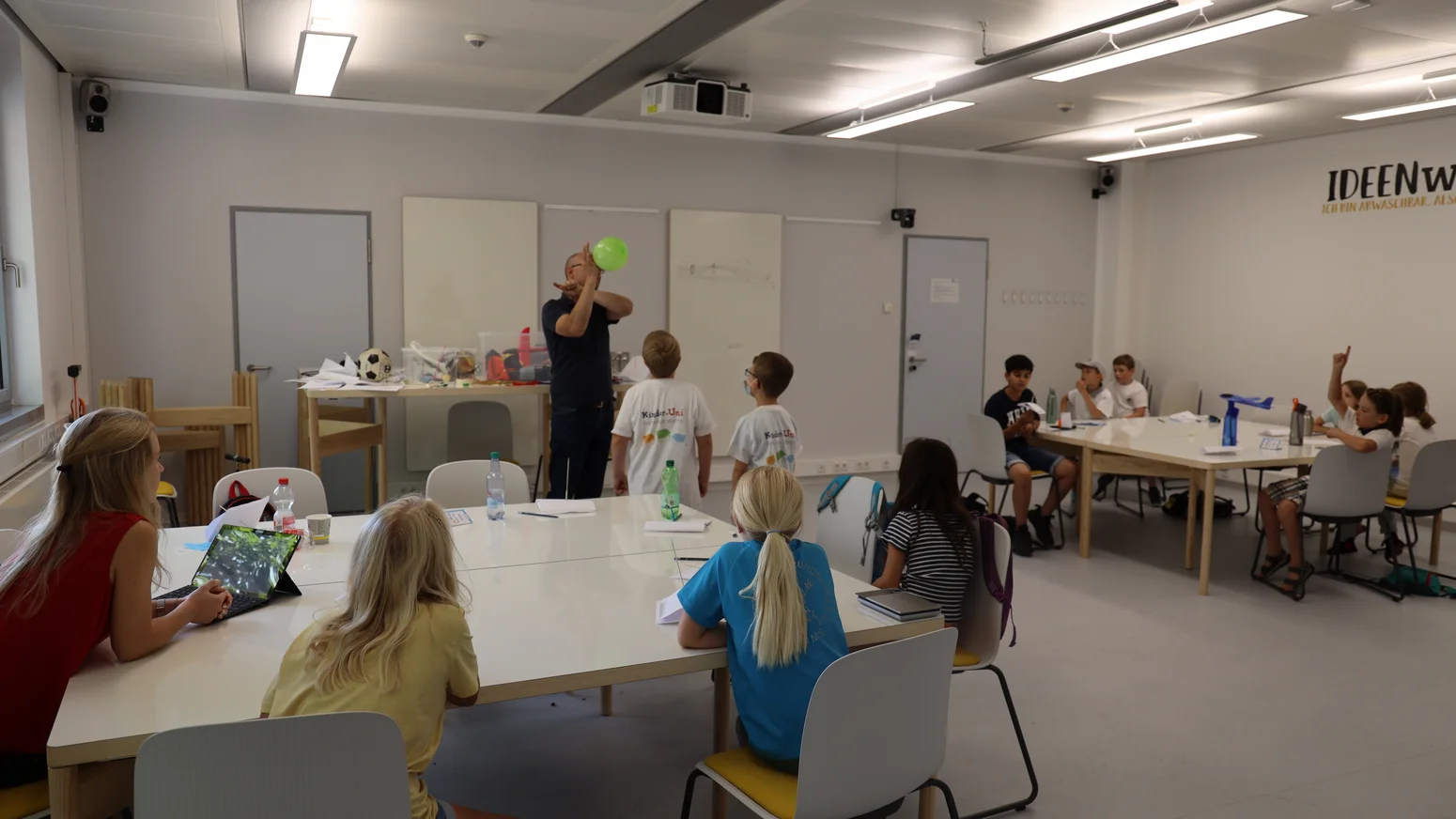 Ein Betreuer wirft einen grünen Luftballon in die Luft vor einer Gruppe von Kindern, die an weißen Tischen sitzen, im Hintergrund der Schriftzug „IDEENWERKSTATT“ an der Wand.
